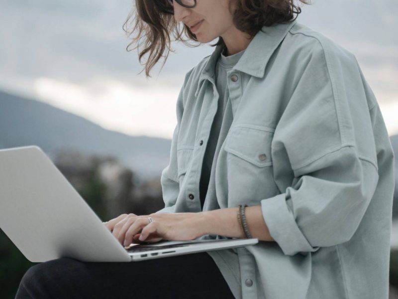 Beautiful curly young girl with glasses working on a laptop with a city view. Modern technologies and urban lifestyle.